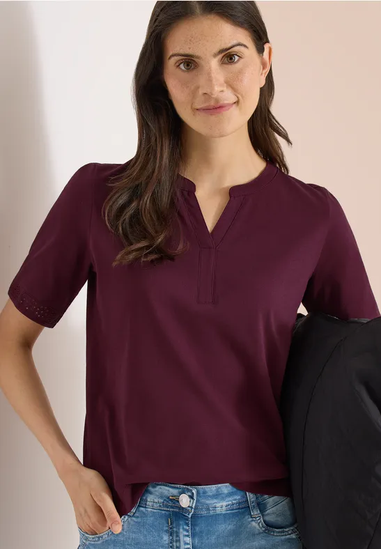 Eine Frau trägt eine bordeauxrote Bluse und Jeans, hält eine schwarze Jacke, Gesicht unscharf.