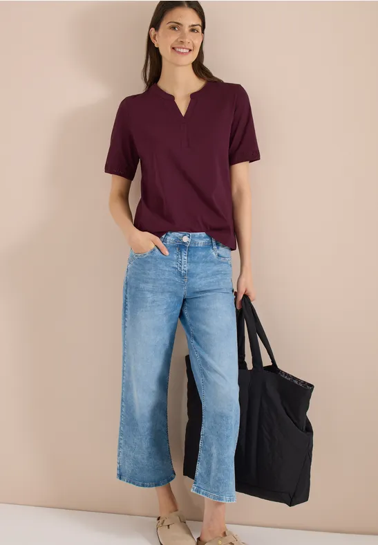 Frau mit burgundfarbenem T-Shirt, weiten Culotte-Jeans und schwarzer Tasche vor beigem Hintergrund.