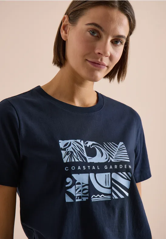 Person trägt ein dunkelblaues T-Shirt mit 'COASTAL GARDEN'-Schriftzug und abstrakten Meeres- und Pflanzenmustern.