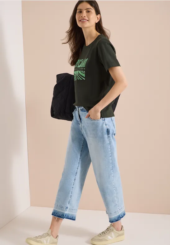 Seitlich stehende Frau in minimalistischem Studio, mit schwarzem T-Shirt mit grünem Print, weiten hellblauen Jeans und großer Tasche.