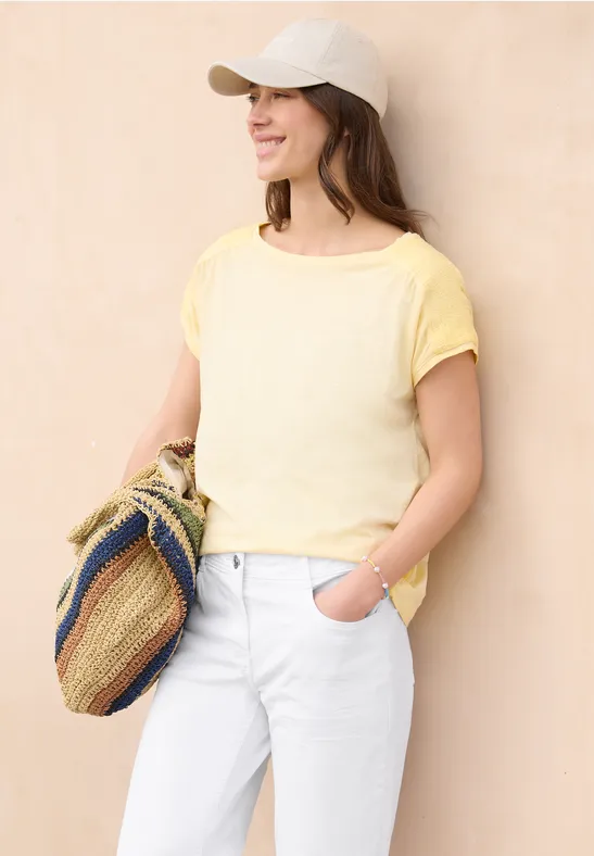 Frau mit gelbem T-Shirt, weißen Jeans, weißer Kappe und Korbtasche vor hellem Hintergrund.