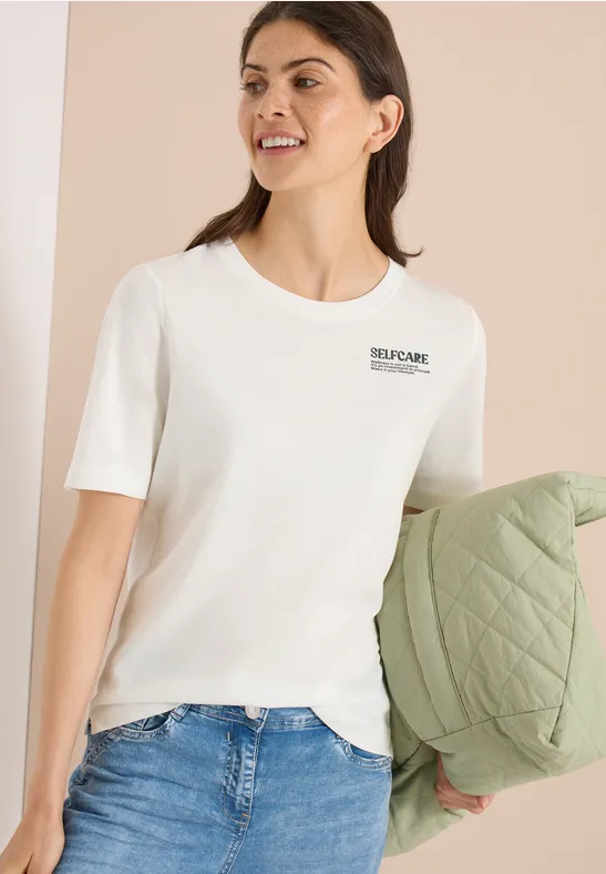 Frau trägt ein weißes, kurzärmeliges T-Shirt und hält eine grüne, gesteppte Jacke in der Hand.