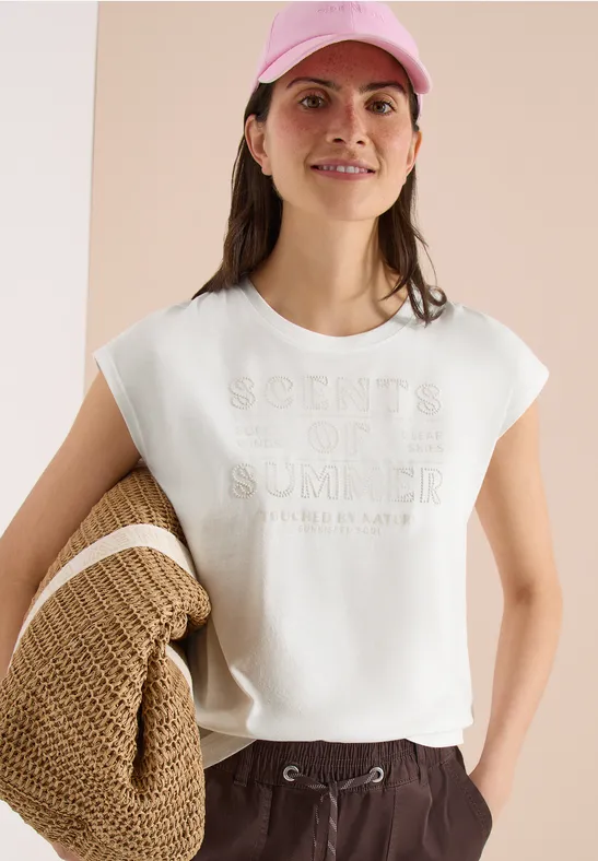 Frau in weißem T-Shirt mit 'Scents of Summer'-Print, rosa Kappe und Strohtasche.