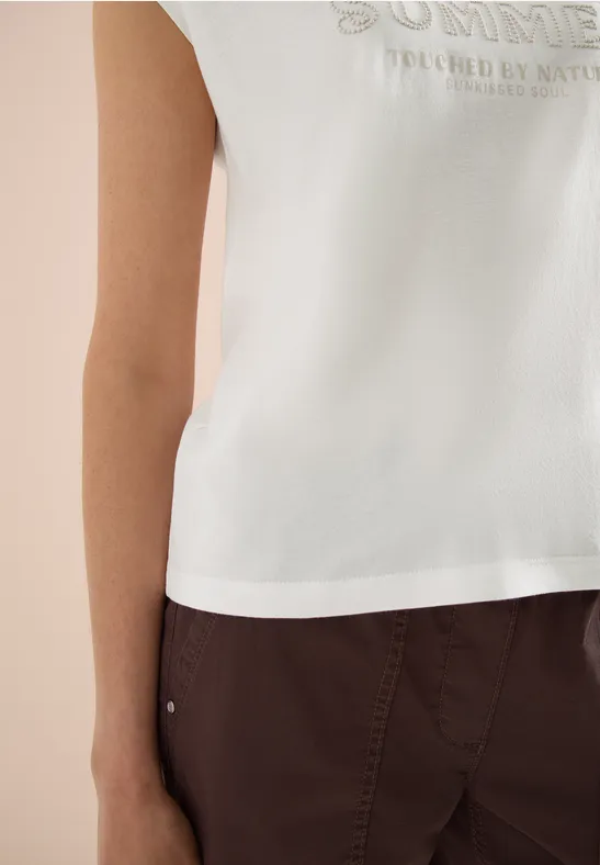 Person trägt ein weißes, ärmelloses Top mit Aufdruck und eine dunkelbraune Hose, Oberkörper abgebildet