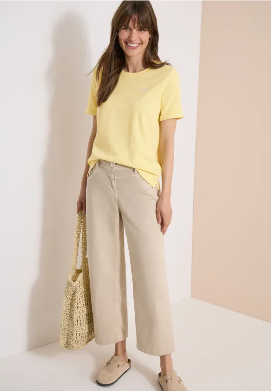 Femme sur fond clair porte un t-shirt jaune, un pantalon beige ample, des chaussures claires et tient un sac filet.
