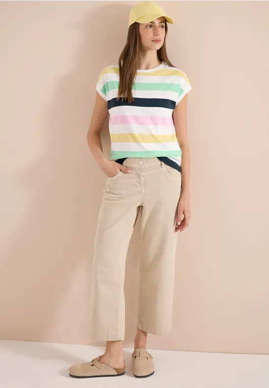 Eine Person trägt bunt gestreiftes T-Shirt, beige Culotte-Hosen und Slipper vor hellem Hintergrund.