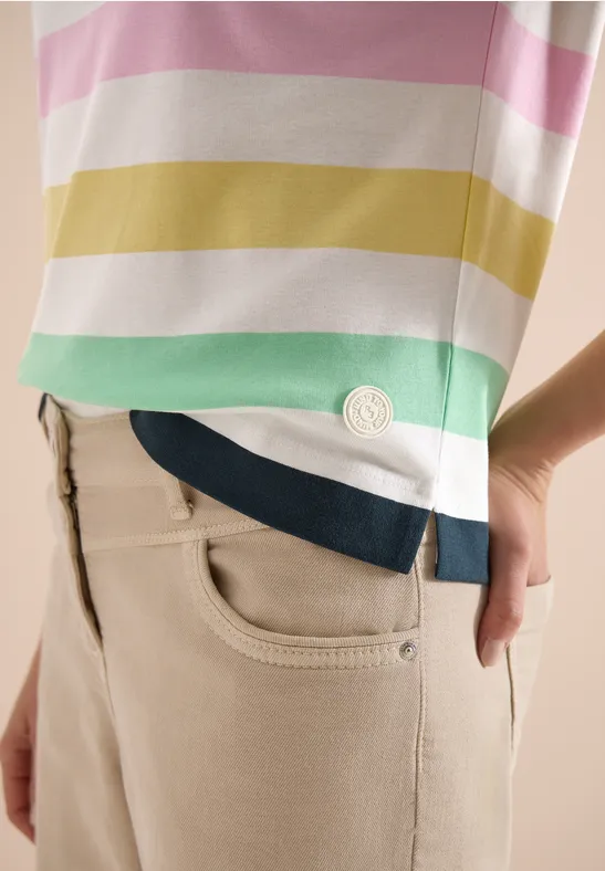 Eine Person trägt beige Hose und ein T-Shirt mit pastellfarbenen Querstreifen und Logo am Saum.