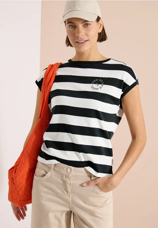Model trägt schwarz-weiß gestreiftes T-Shirt, beige Hose, orange Tasche und Cap.