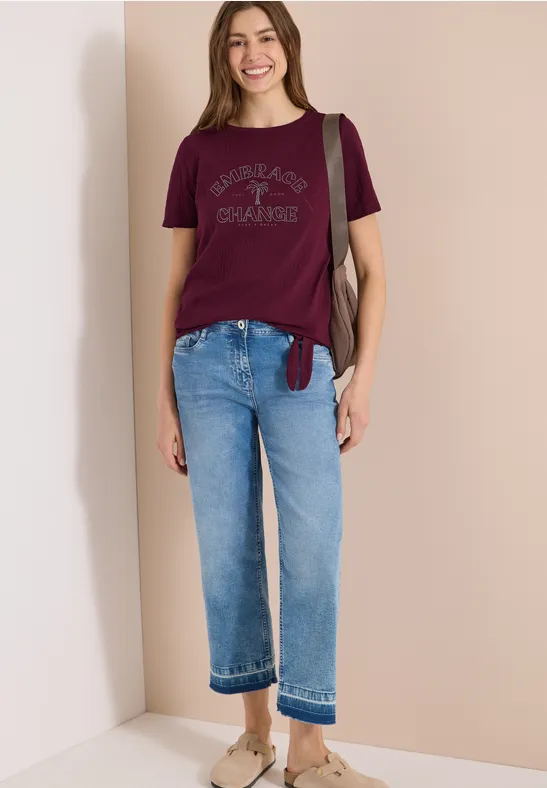 Frau trägt Bordeaux-T-Shirt, hellblaue Jeans und Beige-Slipper, steht vor beigem Hintergrund mit Schultertasche.