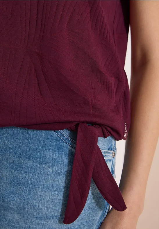 Strukturiertes, pflaumenfarbenes Damen-Top, seitlich geknotet, kombiniert mit blauen Jeans.