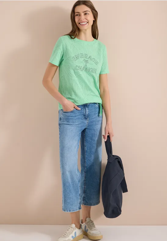 Eine Person mit hellem grünen Print-T-Shirt, weiten blauen Jeans und Sneakern, hält eine dunkle Jacke.
