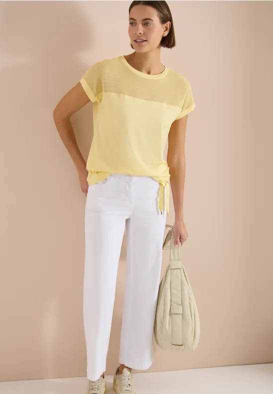 Model trägt transparentes gelbes Kurzarmshirt mit Knotensaum, weiße Cropped-Hose und pastellgrüne Tasche.