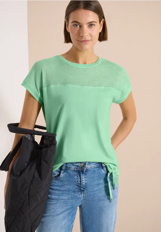 Modell in hellgrünem T-Shirt mit Knotendetail, blauer Jeans und schwarzer Stepp-Tasche.