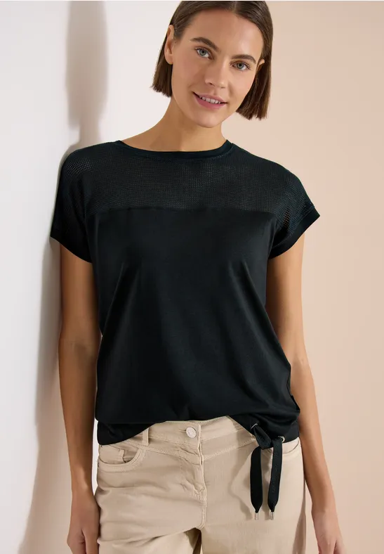 Frau lehnt an Wand, trägt schwarzes T-Shirt mit seitlicher Schleife und heller Hose.
