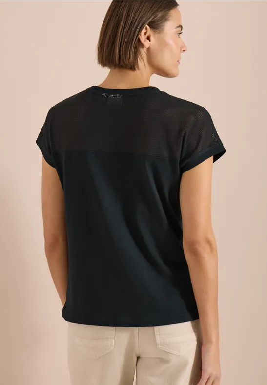 Rückansicht einer Frau in schwarzem T-Shirt mit transparentem Rückenbereich, helle Hose.