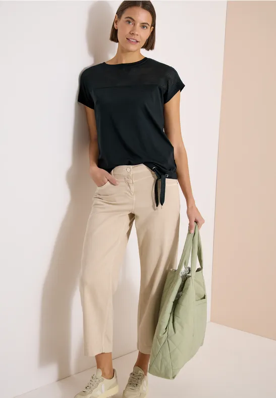 Model trägt schwarzes T-Shirt mit kurzem Arm, beige Hose und hält eine grüne Tasche, steht an Wand.