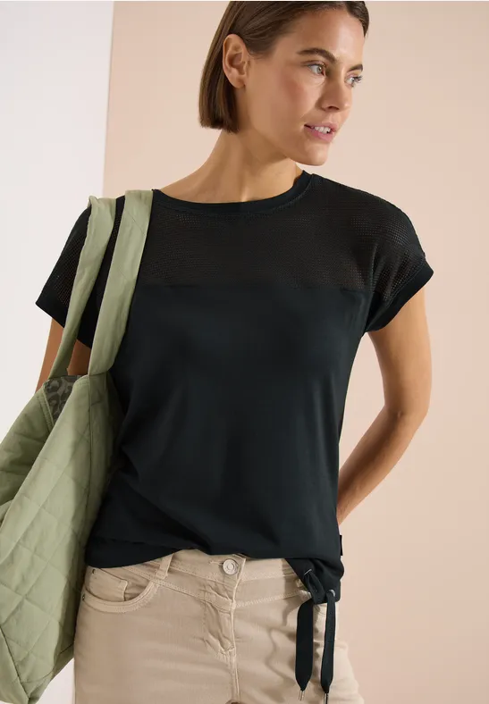 Frau trägt dunkelblaues T-Shirt mit transparentem Oberteil-Einsatz, Saumknoten und grüner Tasche.
