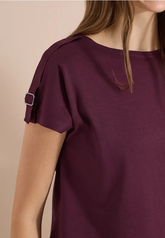 Une femme porte un top manches courtes couleur prune, avec une petite bride et boucle sur la manche.