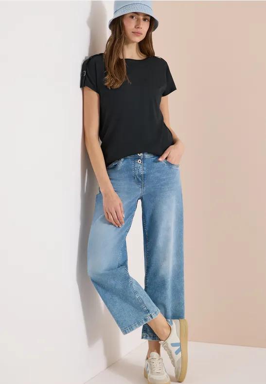 Femme en t-shirt noir, jean large bleu et baskets blanches devant un mur bicolore.