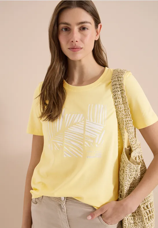 Femme portant un t-shirt jaune avec imprimé graphique blanc, un pantalon beige et un sac crocheté à l'épaule.