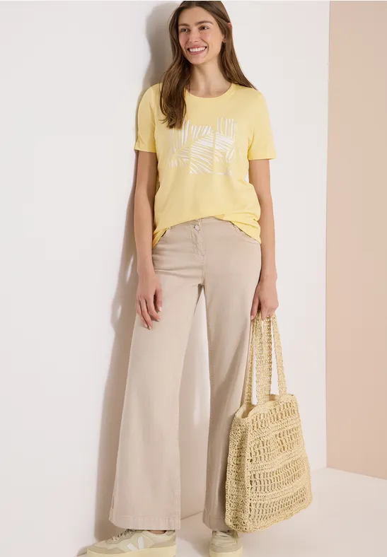 Personne porte un t-shirt jaune, un pantalon large beige et tient un sac de shopping en crochet.