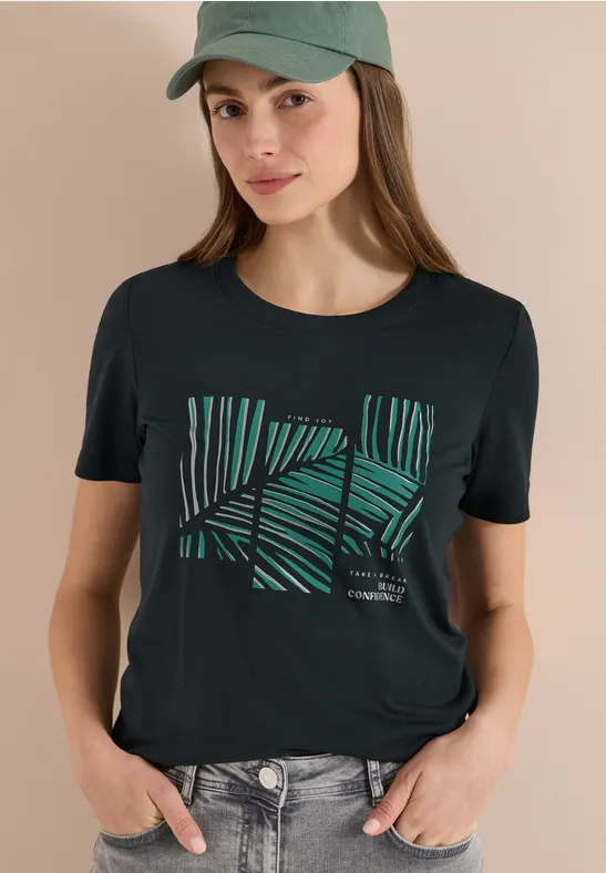Frau mit unscharfem Gesicht trägt schwarzen T-Shirt mit grünem Palmenmuster und Cap.