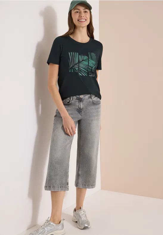 Frau trägt schwarzes T-Shirt mit grünem Muster, graue Culotte-Jeans und Sneaker vor hellem Hintergrund