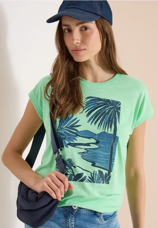 Ein Mädchen trägt grünes T-Shirt mit tropischem Blatt-Print, blaue Kappe und Umhängetasche.