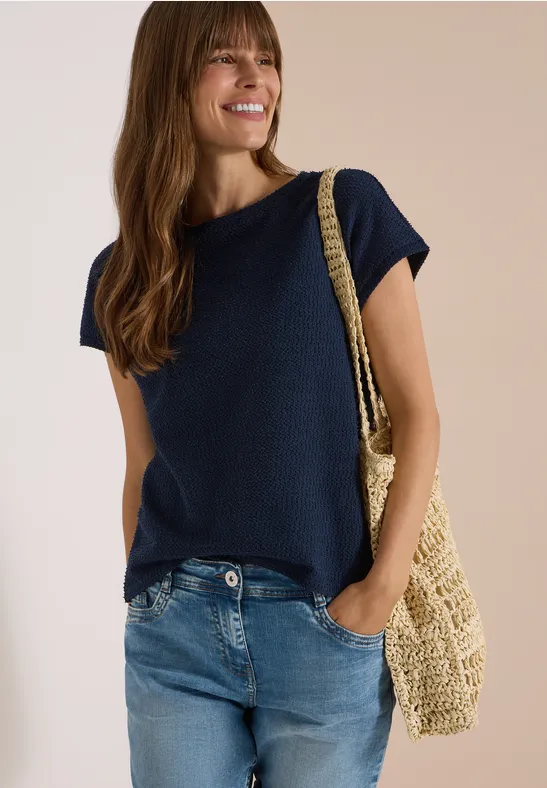 Frau in lässigem kurzärmeligem Shirt, Jeans und Strohtasche, die seitlich lächelt.