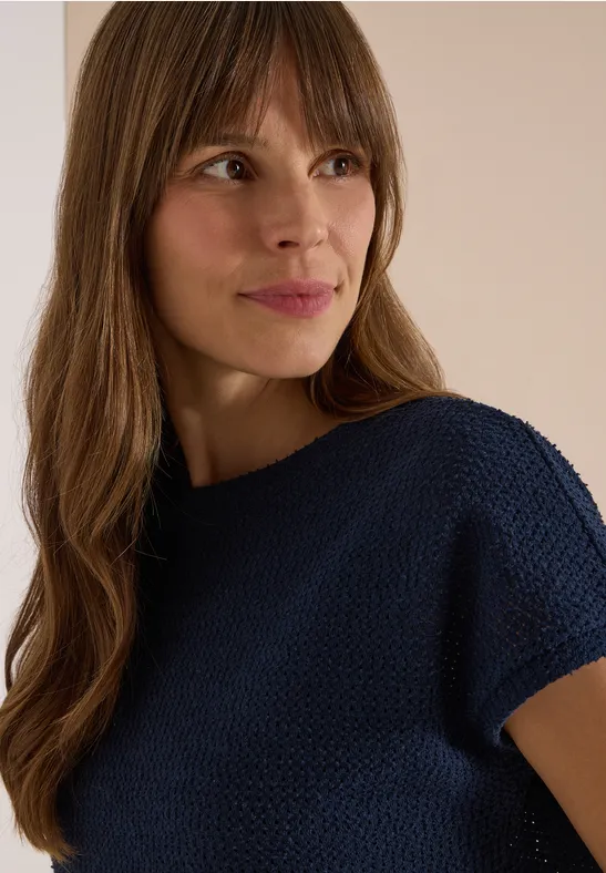 Porträt einer Frau mit langem braunem Haar und dunkelblauem Pullover, die seitlich lächelt.