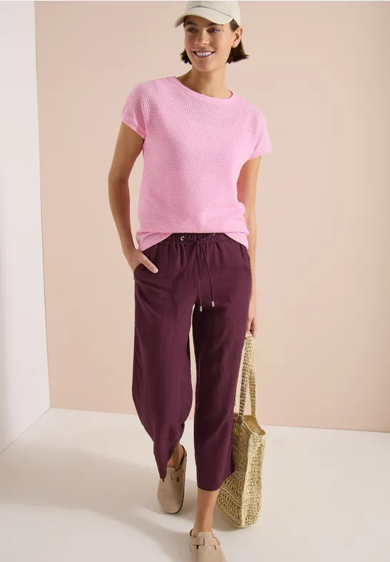 Personne en t-shirt rose, pantalon violet raccourci, sac en paille et chaussures beiges sur fond neutre.