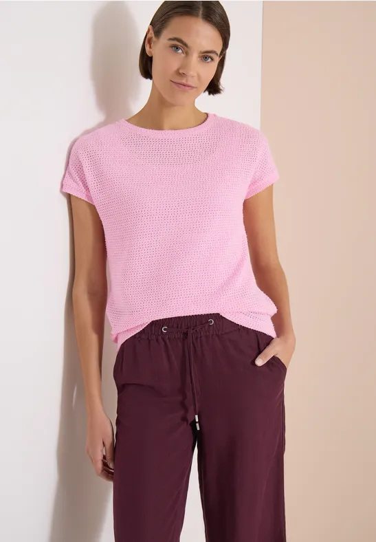 Femme devant un mur clair, porte un t-shirt rose et un pantalon ample bordeaux.