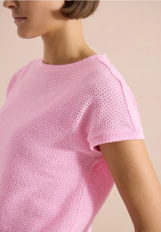 Une personne porte un pull rose à manches courtes en maille sur fond clair.
