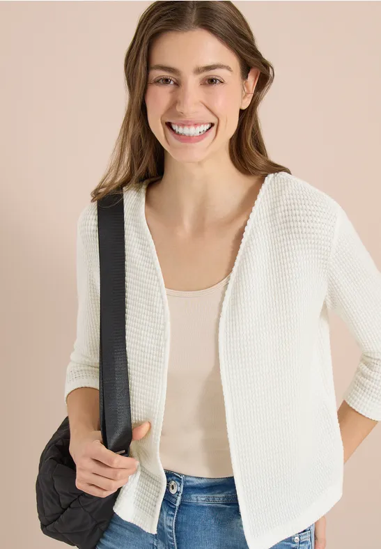 Femme porte un gilet blanc, débardeur beige et jeans, tient une bandoulière noire