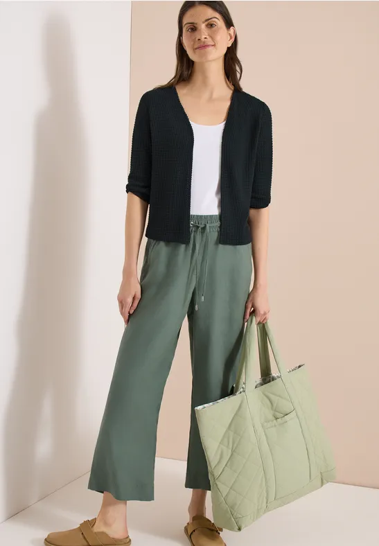 Femme en débardeur blanc, gilet noir et pantalon ample vert, tenant un grand sac matelassé vert clair.