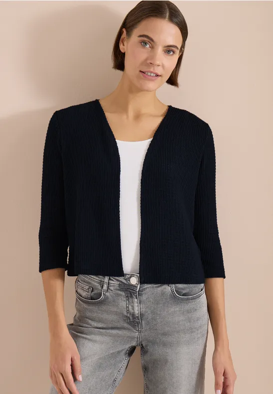 Frau trägt kurze schwarze Strickjacke, weißes Top, graue Jeans vor beigem Hintergrund.