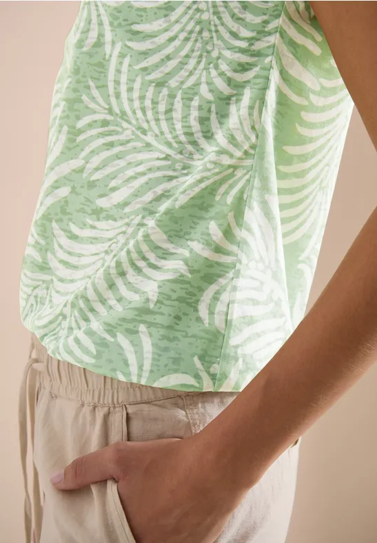 Nahaufnahme eines Oberkörpers in beiger Hose, grünes ärmelloses Top mit weißem Palmenblatt-Print, Hand in Tasche.