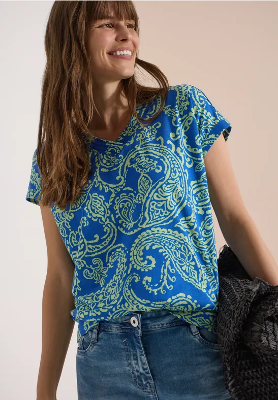 Frau trägt blaues Paisley-T-Shirt, Jeans und hält eine schwarze Tasche vor neutralem Hintergrund.