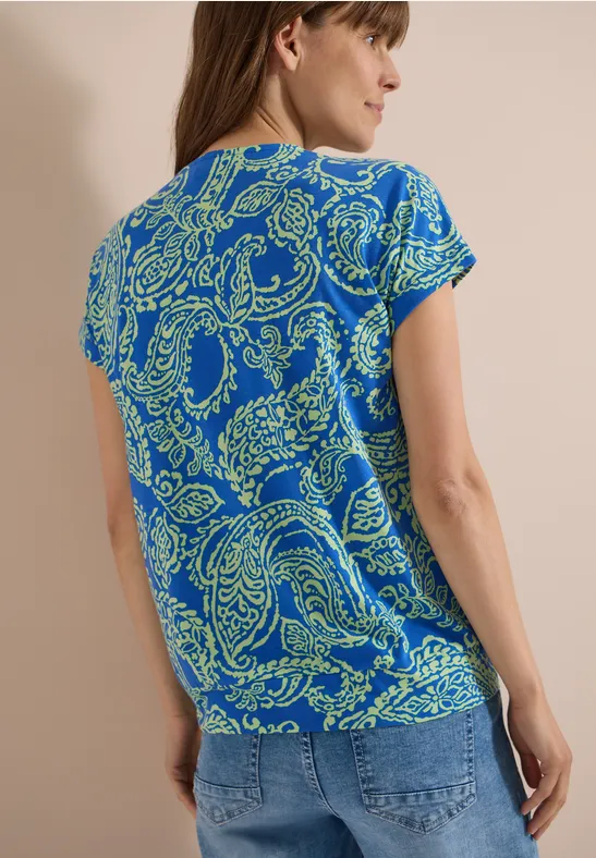 Frau von hinten mit blauem T-Shirt und grünem Paisleymuster, dazu Jeans.