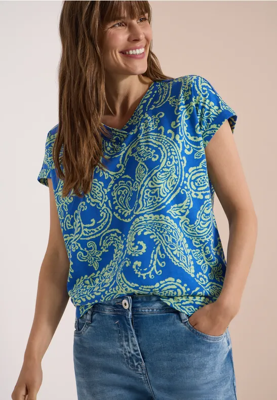 Frau trägt blaues T-Shirt mit Paisley-Muster und Jeans, Hand in Tasche