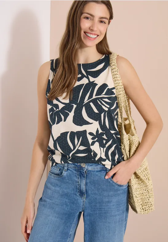 Vrouw in wit topje met zwart bladmotief, blauwe jeans en gevlochten schoudertas.