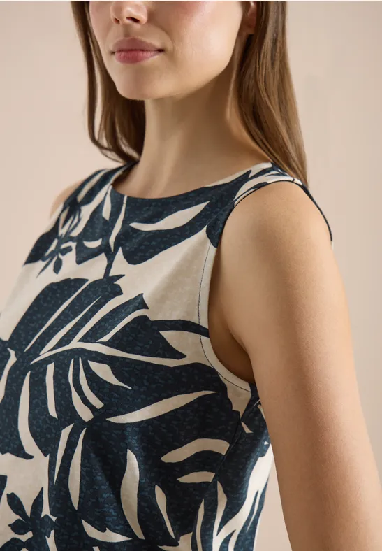 Close-up van een vrouw in een mouwloze top met donkerbladerprint op lichte achtergrond.