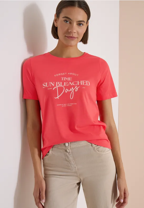 Frau im roten T-Shirt mit Aufschrift und beigen Hosen vor hellem Hintergrund.