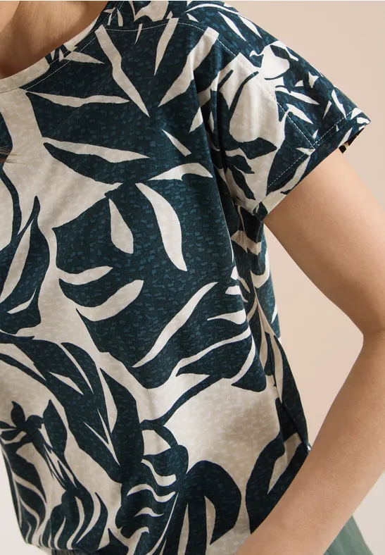 Nahaufnahme eines beigen T-Shirts mit dunkelblauem, abstraktem Blattmuster auf Schulter und Arm.