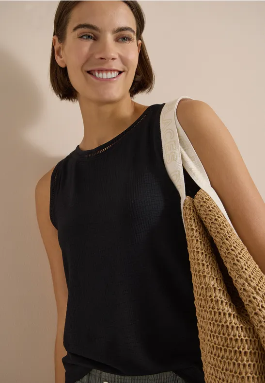 Femme en débardeur noir portant un grand sac en paille tressée sur fond neutre.