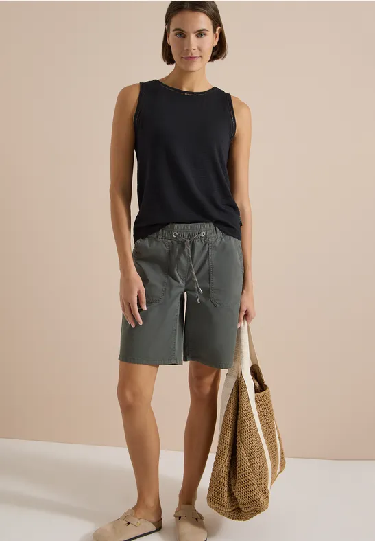 Femme en débardeur noir, short gris et sandales, portant un sac de plage en osier.
