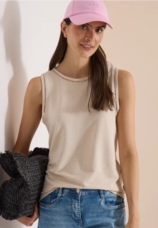 Frau in beigem Tanktop, blauer Jeans, rosa Cap, mit geflochtener Tasche vor hellem Hintergrund.