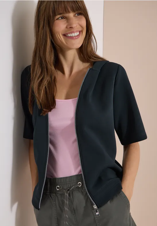 Femme appuyée contre un mur, cardigan noir sur débardeur rose et pantalon gris.