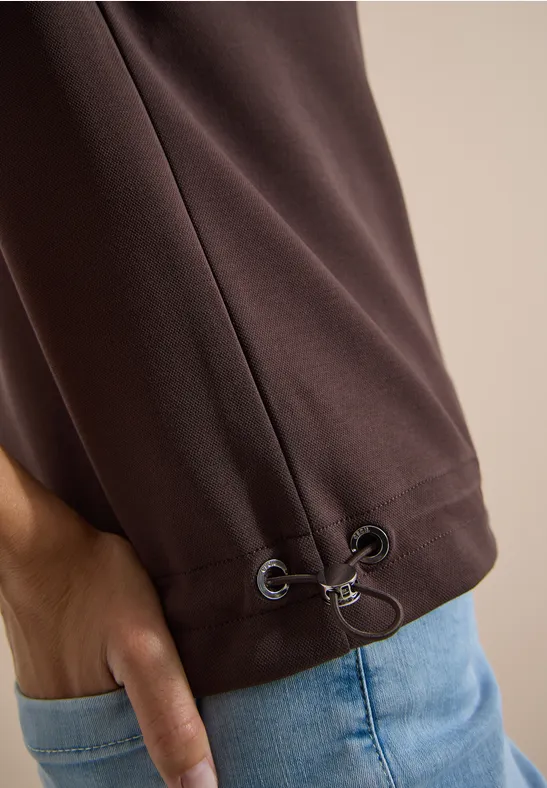 Braunes Oberteil mit Kordelzug am Saum, Hand steckt locker in blauer Jeans.
