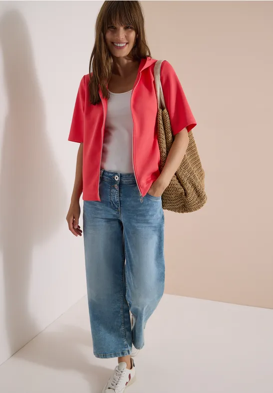 Frau trägt weißes Top, pinke Jacke, weite Jeans, weiße Sneaker und Korbtasche vor hellen Wänden.